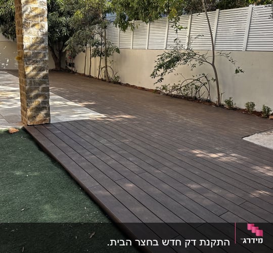 דק עץ בגינה עם עצים וגדר לבנה ברקע